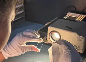 Technician wearing cleanroom gloves inspecting a small optical glass window under a focused projector light in a darkroom to detect microscopic surface defects
